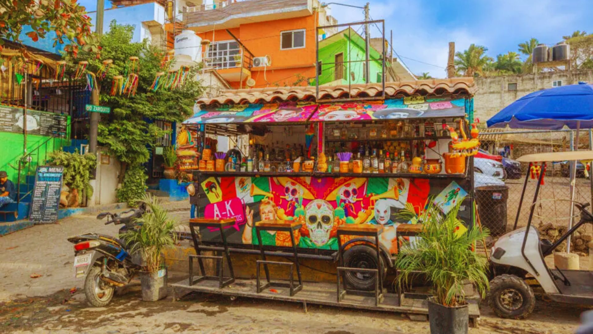 A vibrant street food cart in a colorful neighborhood, adorned with Day of the Dead skull art and surrounded by plants. The scene feels lively and festive.