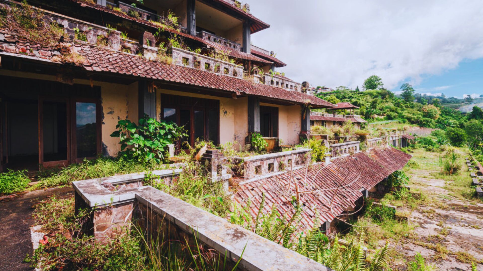 Abandoned resort with crumbling walls and overgrown plants, set in a lush, hilly landscape under a partly cloudy sky, conveying a sense of decay.