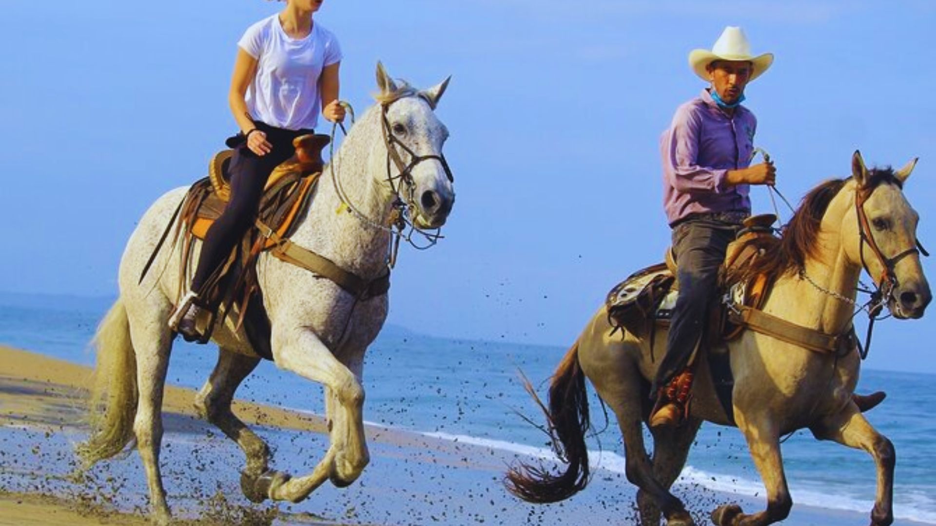 Two people ride horses on a sandy beach. The woman on a white horse wears a white shirt, the man in a cowboy hat on a brown horse wears a purple shirt. The scene conveys adventure and freedom.