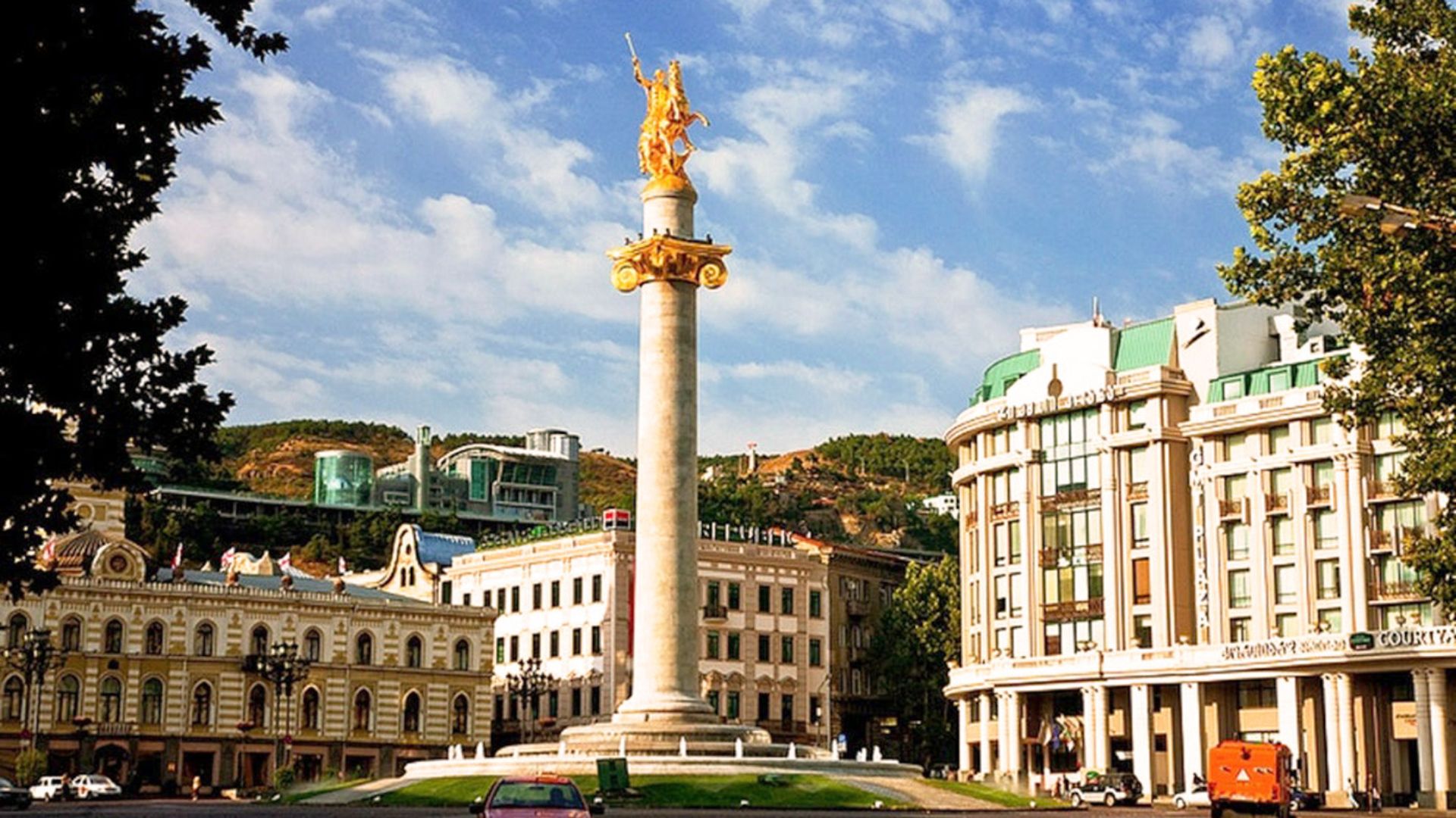 A large white statue stands prominently in the center of a bustling city, surrounded by buildings and pedestrians.