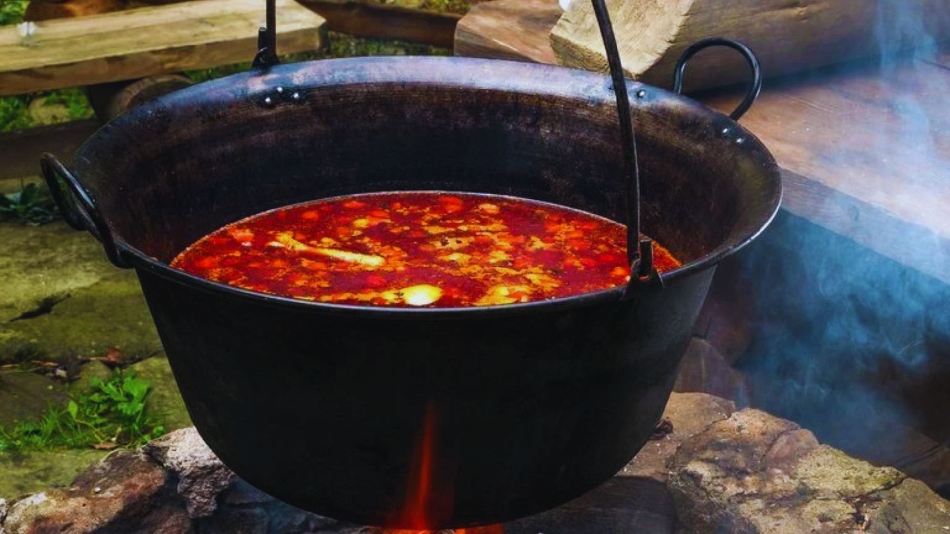 A pot of soup simmers over an open fire, surrounded by glowing embers and a rustic outdoor setting.