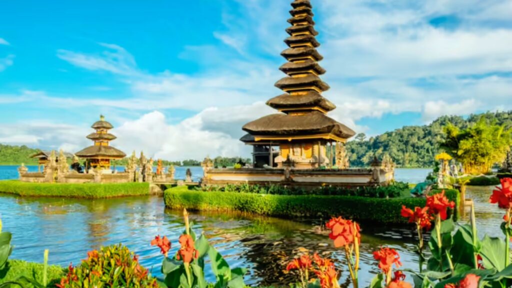 A serene view of Bali's Ulun Danu Beratan Temple surrounded by water, lush greenery, and vibrant red flowers under a bright blue sky.