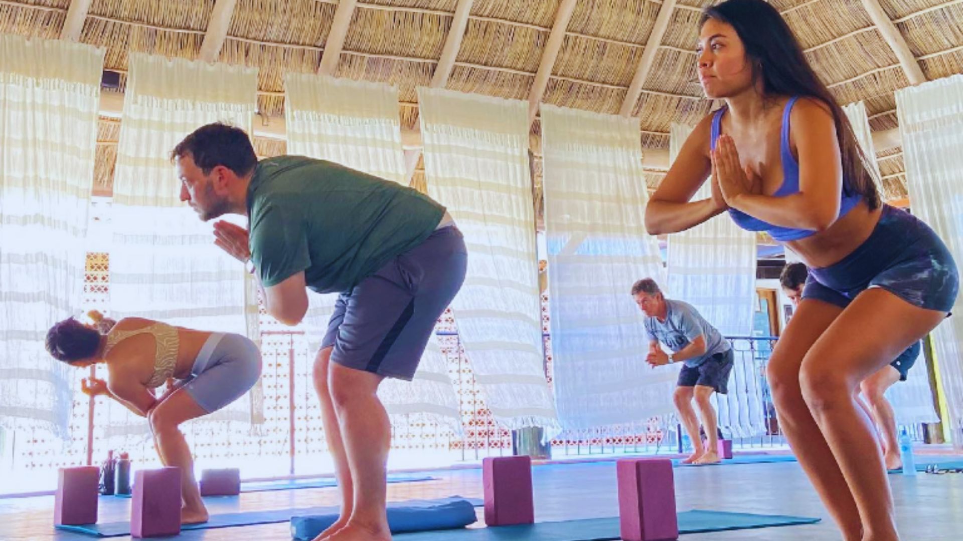 A group of people in a yoga class, practicing chair pose on mats in a sunlit studio with wooden ceiling beams and soft curtains. Focused and calm atmosphere.