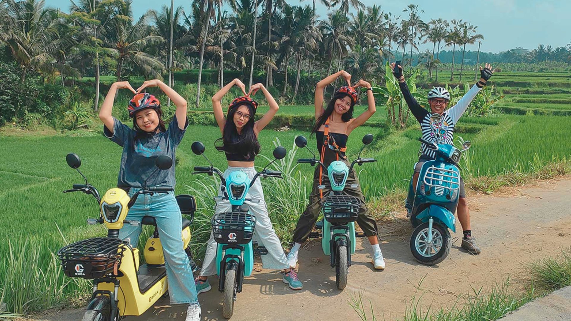 Four people on colorful scooters strike playful poses on a rural path, surrounded by lush green fields and tall palm trees, conveying joy and adventure.