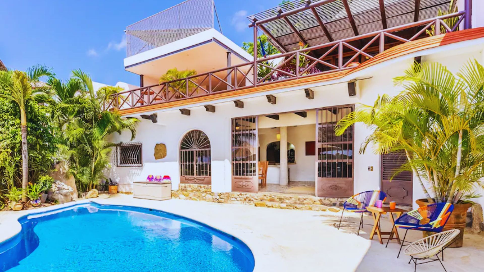 A vibrant villa with white walls, wooden balcony, and lush palm trees surrounding a clear blue pool. Sunlit, inviting outdoor seating area creates a relaxing ambiance.
