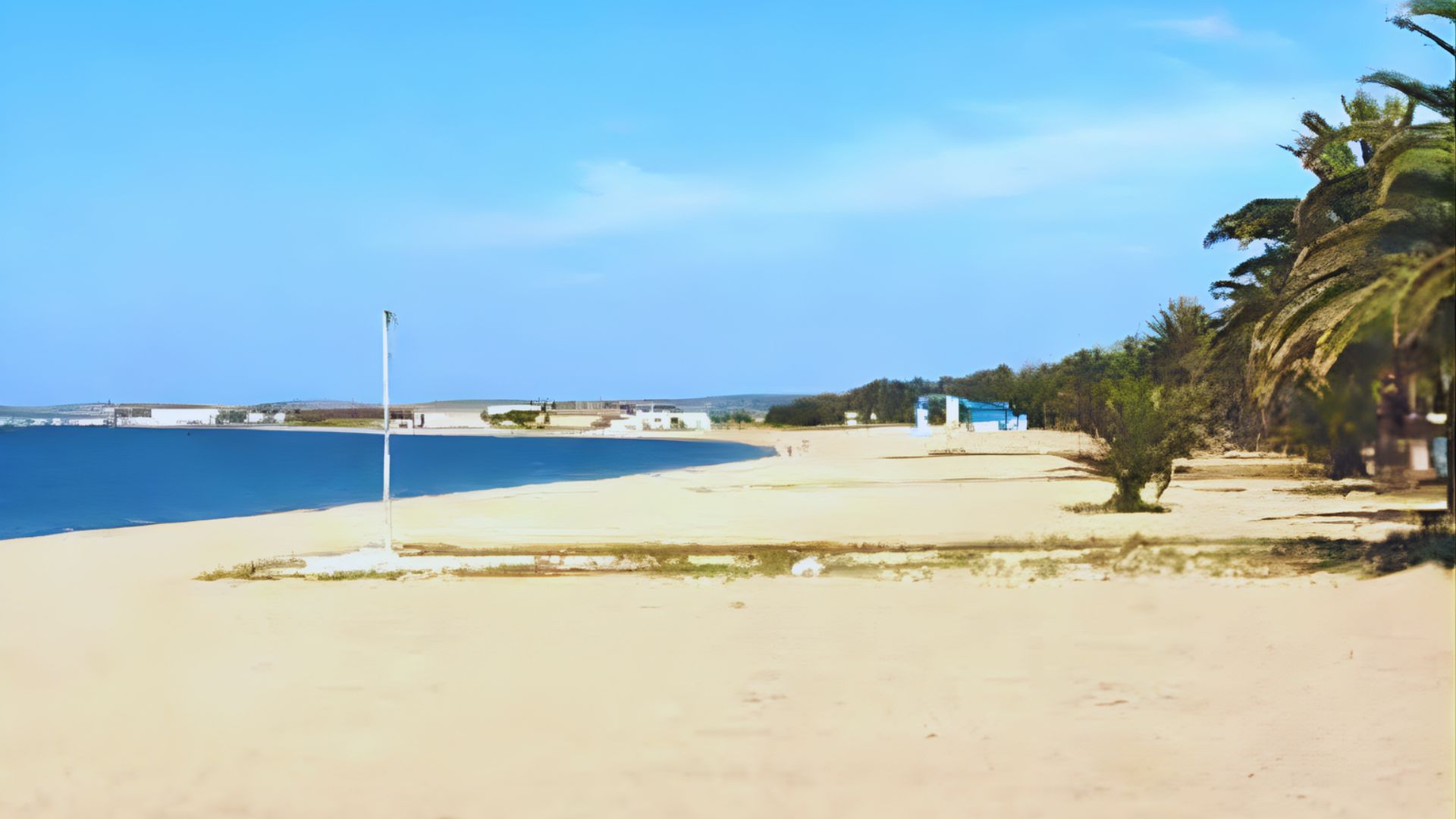 Sandy beach with clear blue sky and calm sea. A few scattered buildings and palm trees line the horizon, creating a serene, tropical atmosphere.
