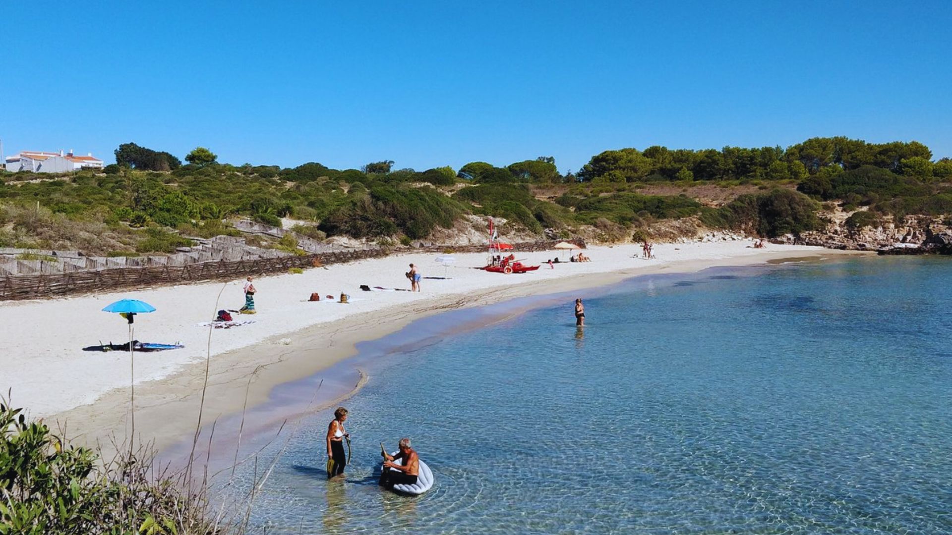 A serene beach scene with clear blue water and a sandy shore. People relax under a bright sun, with some swimming and others sunbathing. Lush greenery frames the beach, creating a tranquil summer atmosphere.