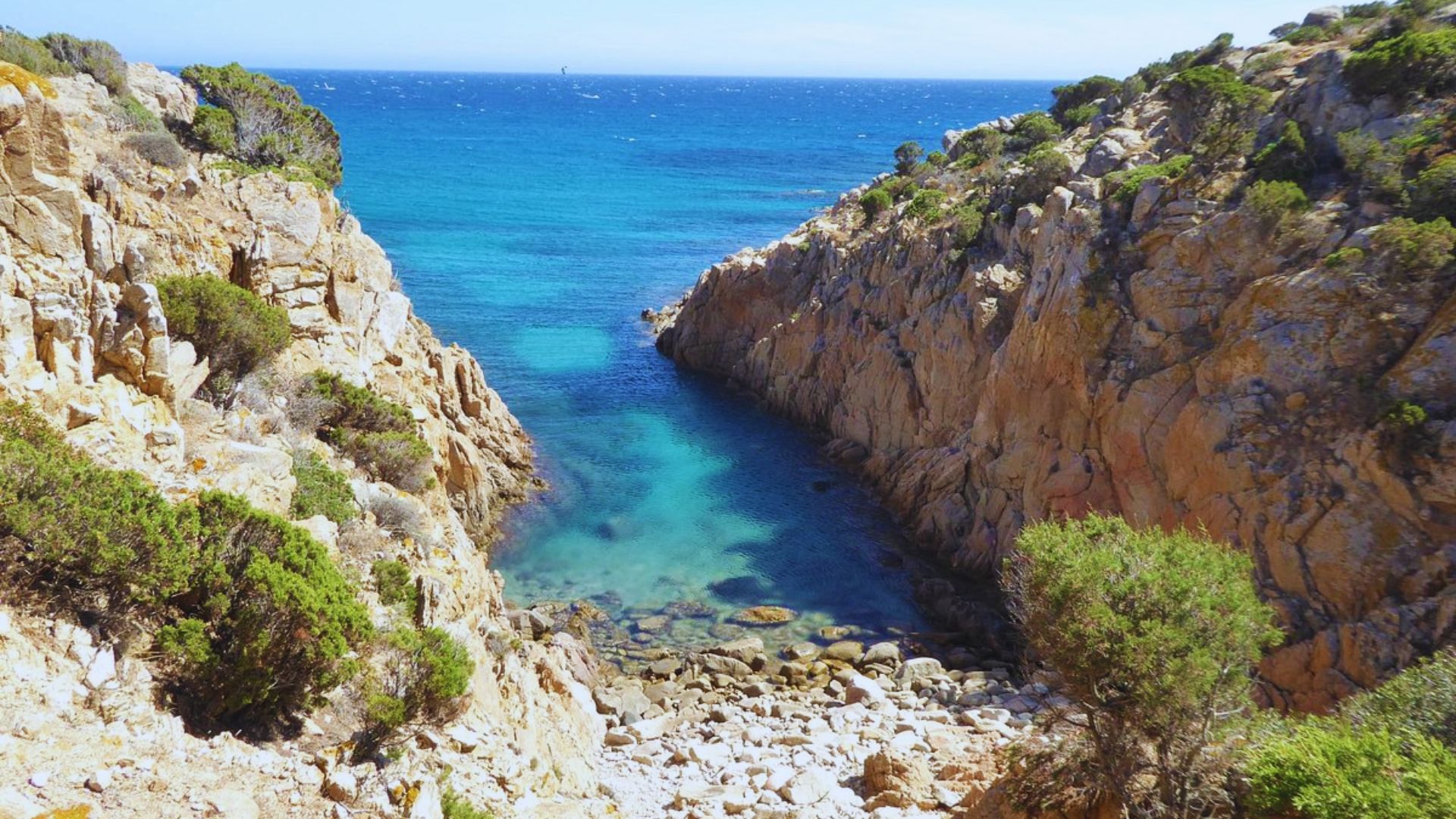 A narrow cove opens to clear turquoise sea, framed by rugged, rocky cliffs with sparse greenery under a bright blue sky, evoking tranquility.