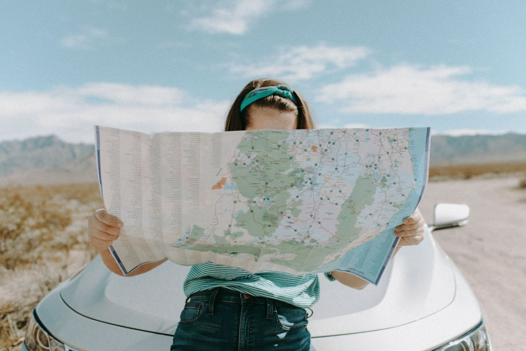 Person with long hair, wearing a striped shirt and headband, reads a map while leaning against a car in a desert under a blue sky.
