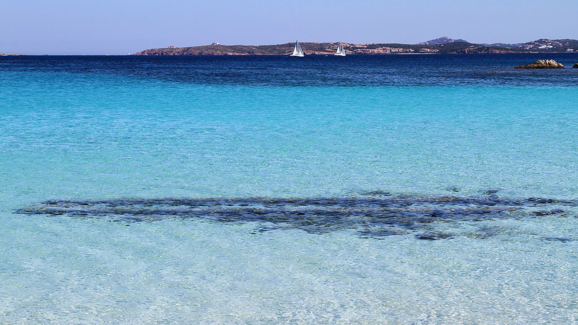 Clear turquoise beach water with darker patches of seaweed in the foreground. Two sailboats are visible on the distant deep blue horizon.