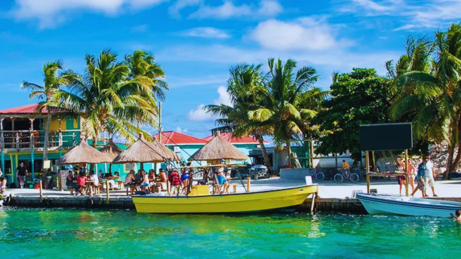 People relax on boats in the water, with a sandy beach visible in the background under a clear blue sky.