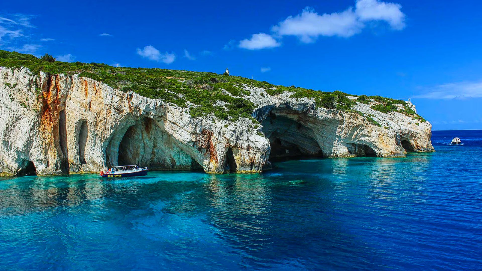 A vibrant blue sea contrasts with rugged white cliffs and green vegetation. A small boat is anchored near the caves, evoking a sense of serene exploration.