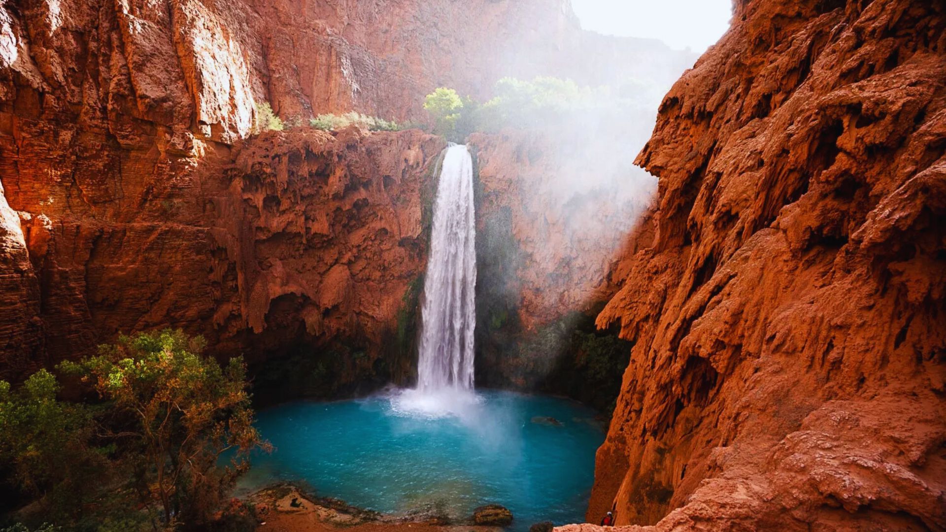 A stunning waterfall cascades into a turquoise pool, nestled between steep red rock cliffs, with lush greenery and sunlight streaming through mist.