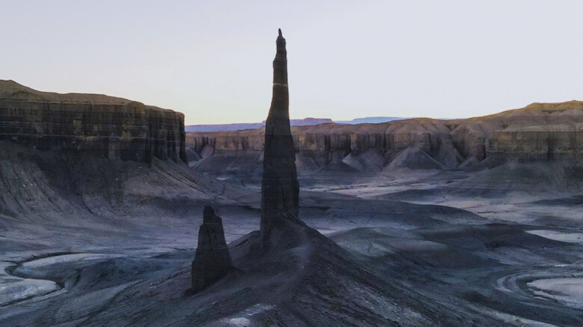 A tall, slender rock formation rises from a rugged, barren landscape at dusk. Cliffs surround the scene, casting long shadows in a calm, desolate atmosphere.