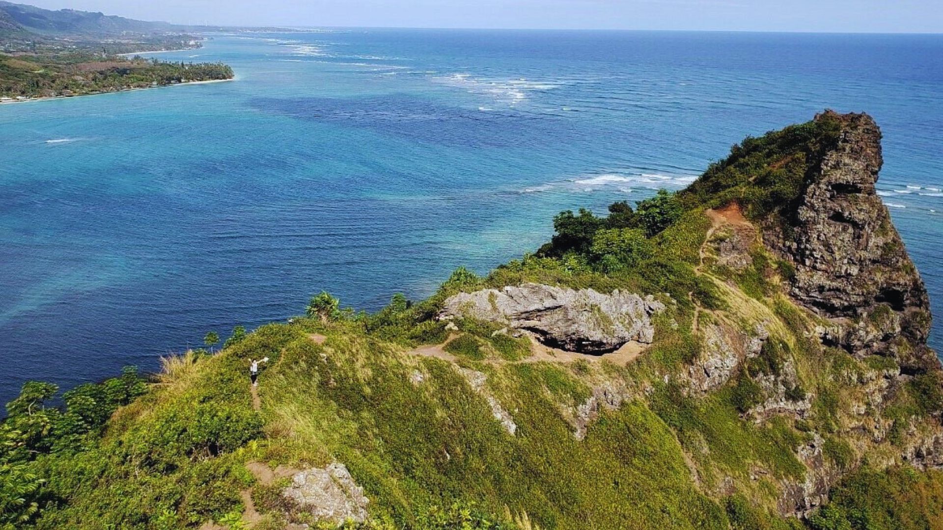 A rugged, green cliff juts into the vibrant blue ocean, with lush vegetation on top. Waves gently crash below, creating a serene and breathtaking coastal scene.