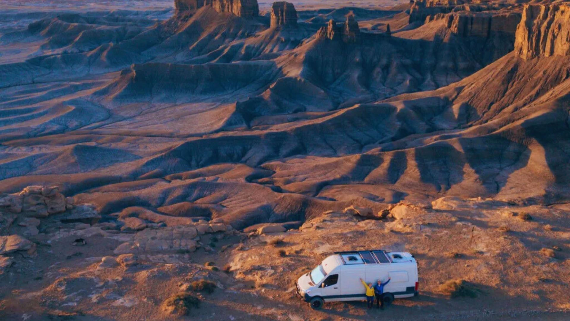 A white camper van parked on a rugged desert landscape at sunset, with towering rock formations in the background, and two people standing nearby.