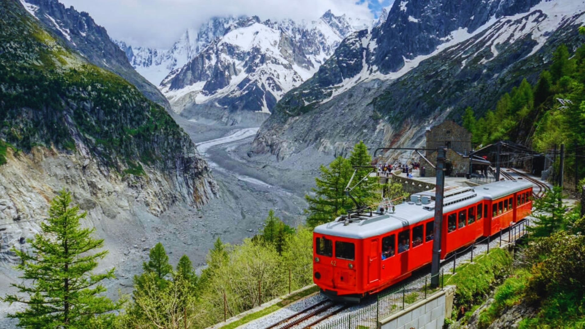 A red train travels through a scenic mountain landscape, surrounded by lush greenery and towering peaks.