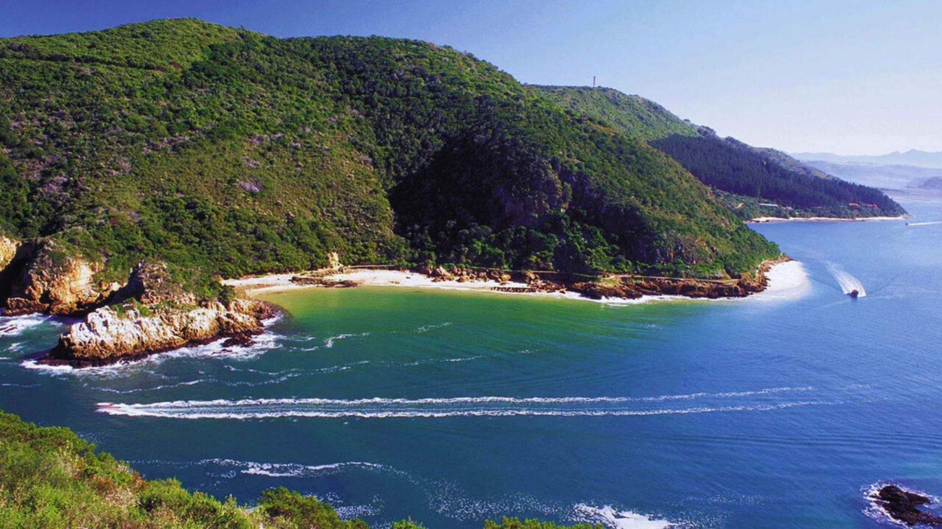 A boat sails along the coast, with a lush green mountain rising in the background.