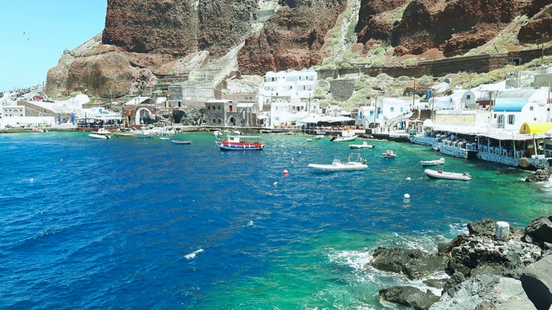 A scenic view of the Santorini harbor in Greece, showcasing boats and whitewashed buildings against a blue sky.