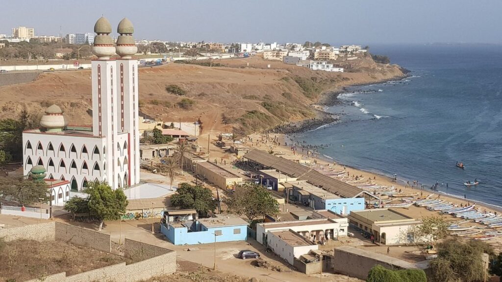A white mosque stands on the beach, overlooking the ocean under a clear blue sky.