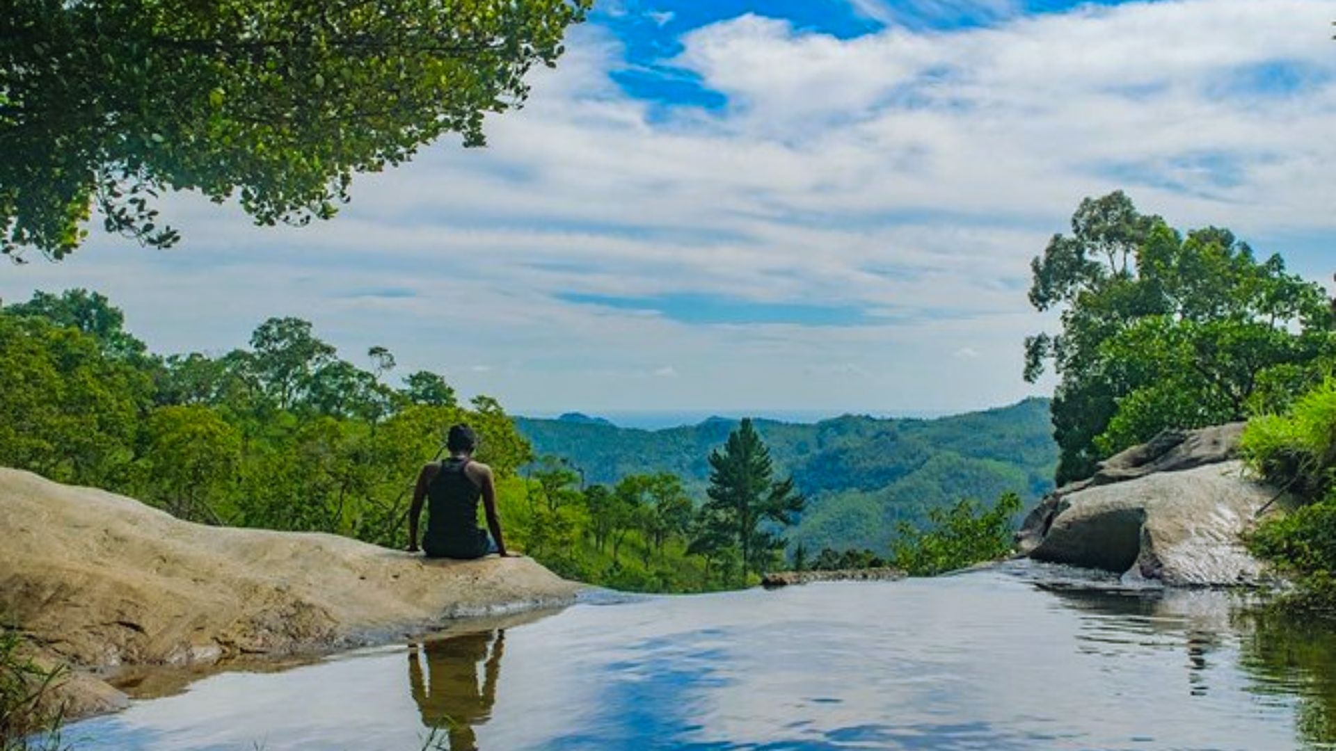 A person sits on a rock, gazing at a flowing river surrounded by lush greenery.