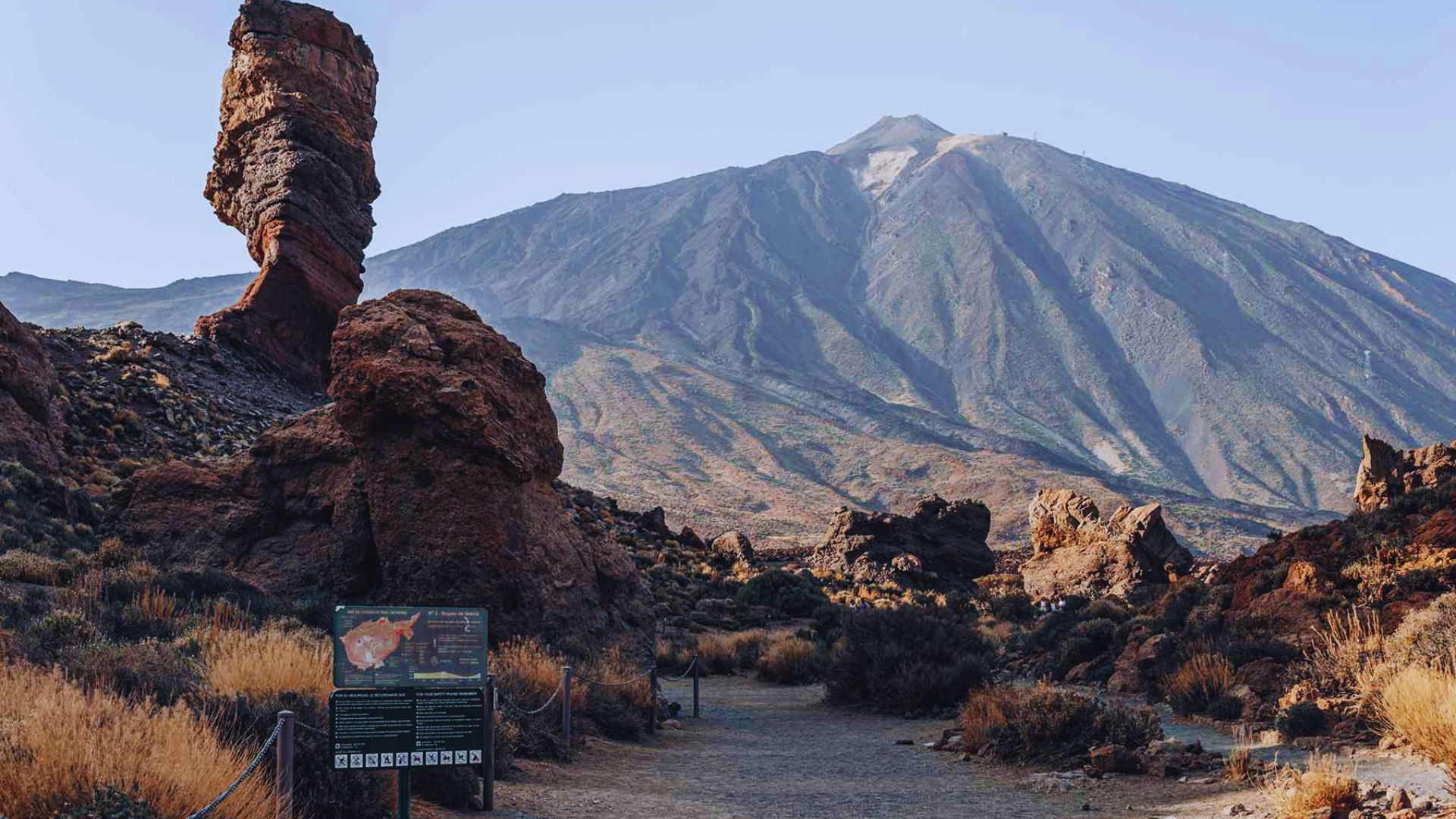 A winding trail leads to a mountain, marked by a wooden sign indicating the path ahead.