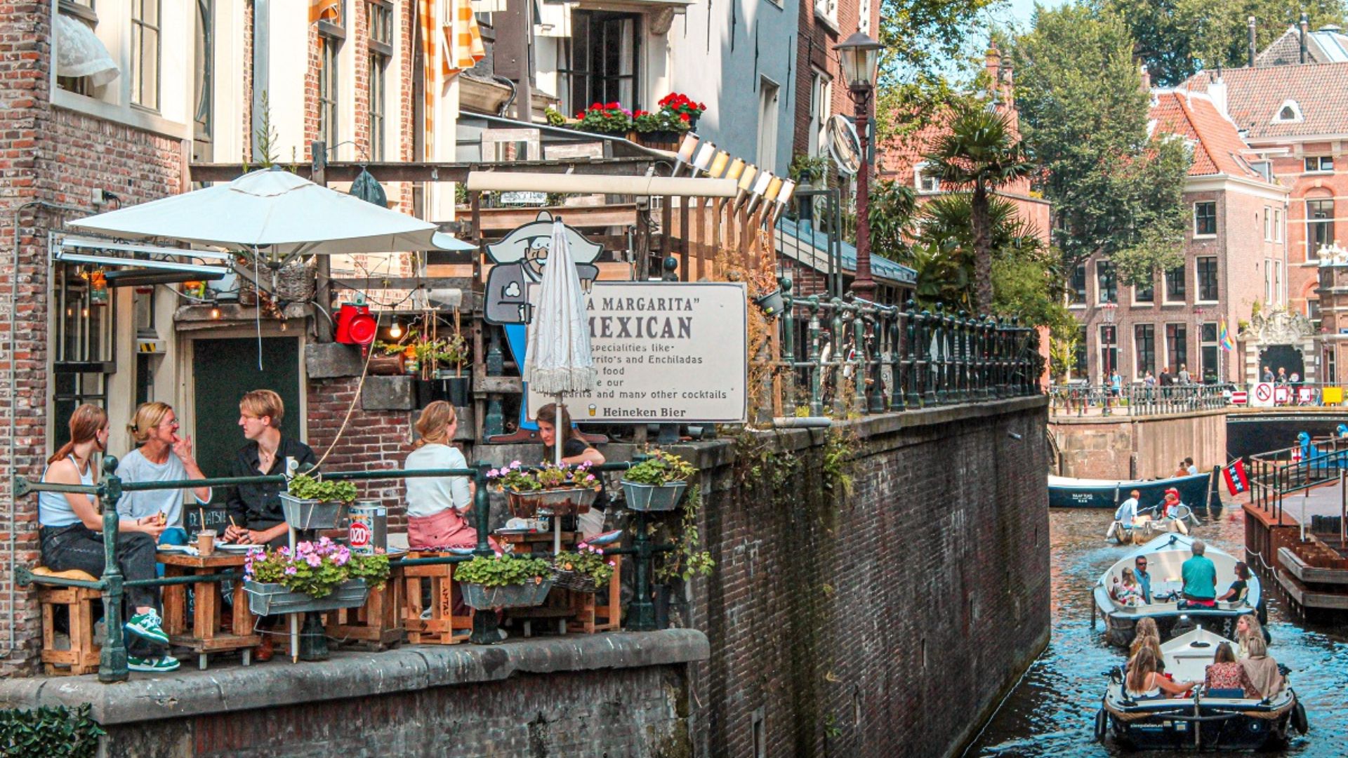 A lively canal scene featuring diners at tables and a boat navigating the waterway.