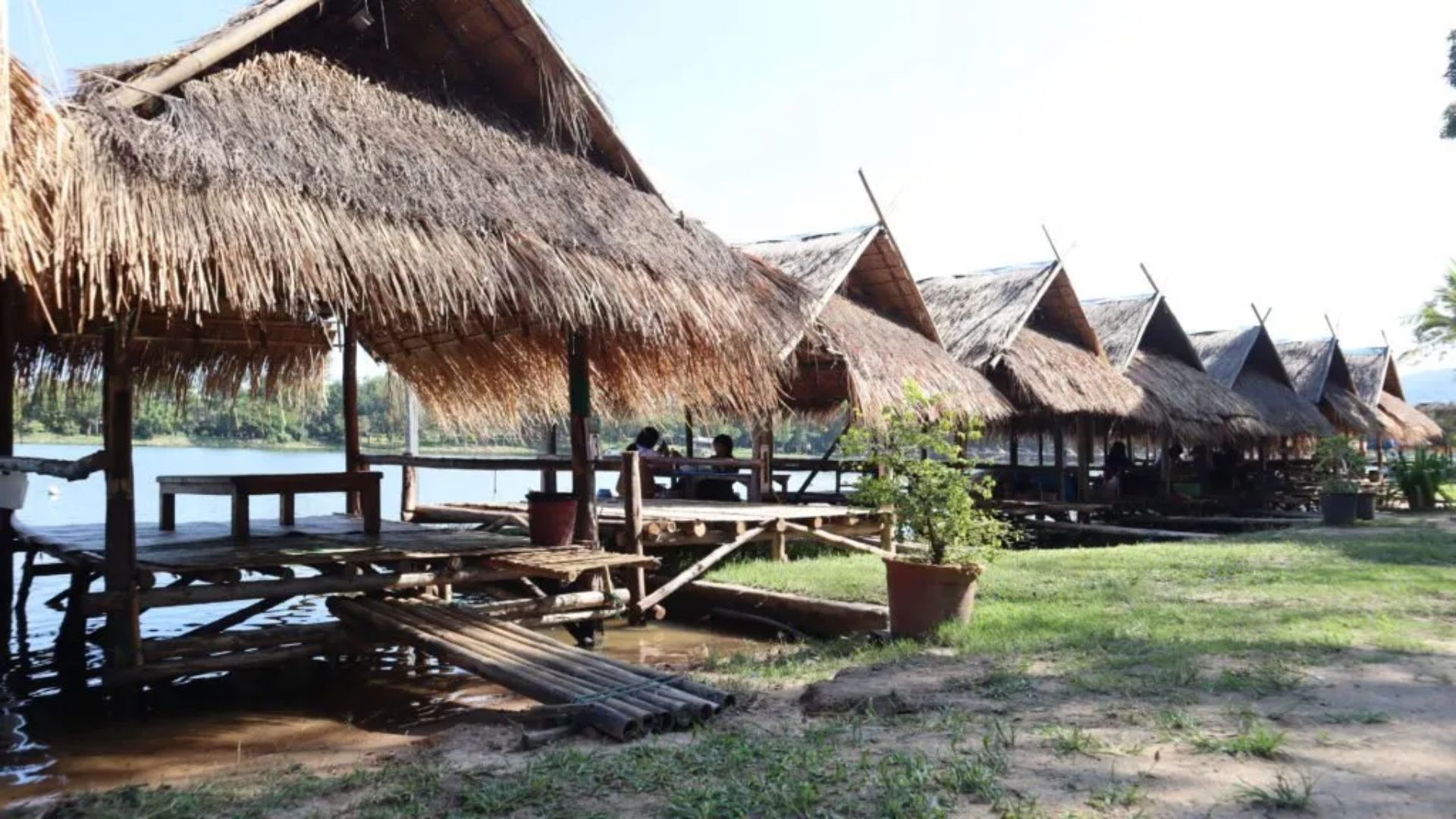 Several thatched huts are positioned along the tranquil shore of a lake, surrounded by lush greenery.