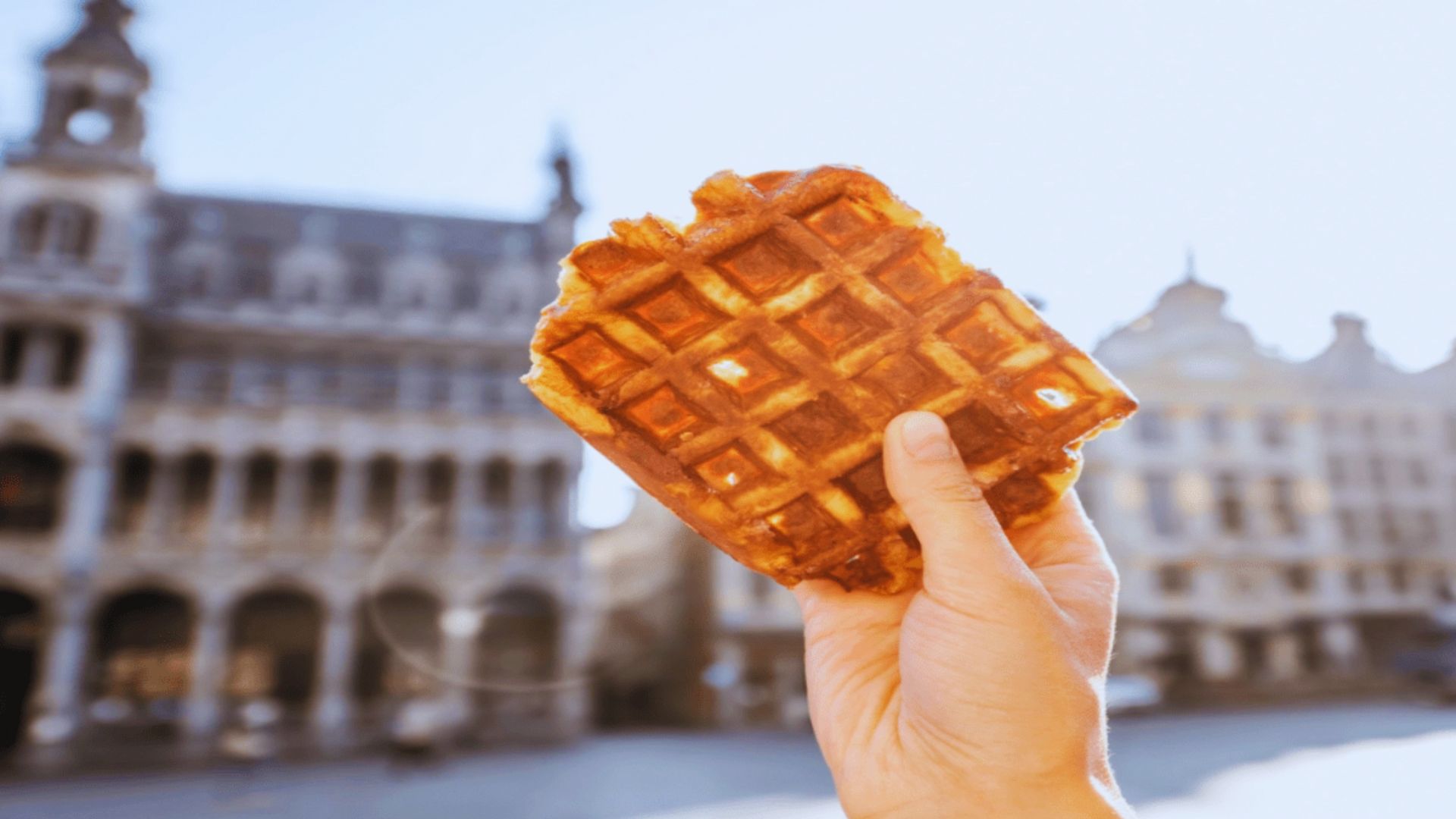 A person holds a waffle in front of a building, showcasing the treat against an urban backdrop.