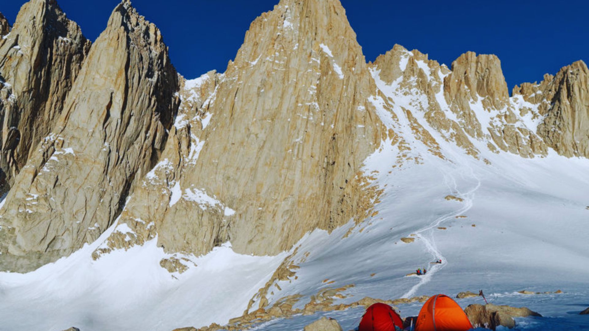 Two tents are pitched on the snow, with a majestic mountain backdrop in a serene winter landscape.