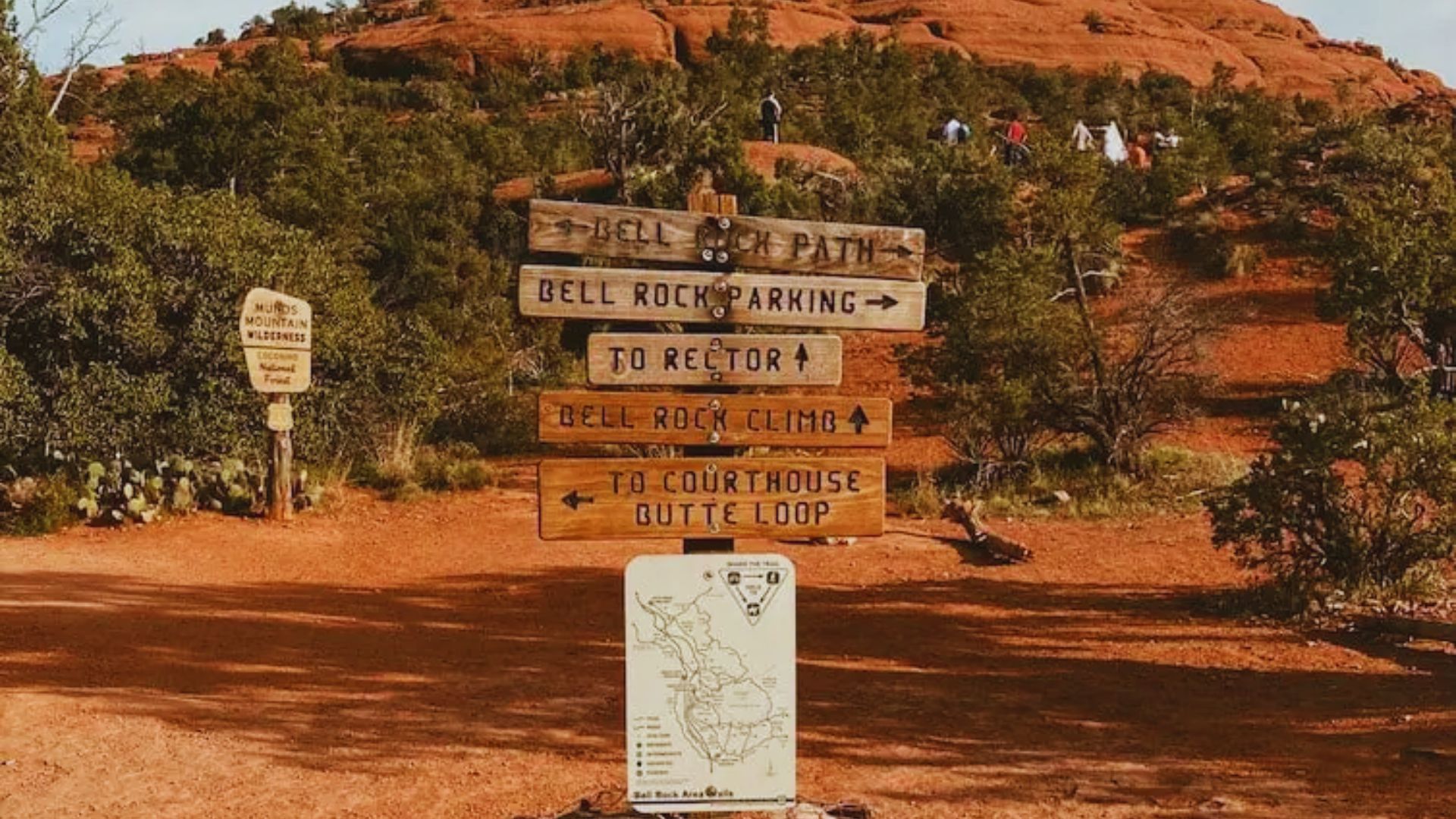 Wooden trail signs point to Bell Rock paths, surrounded by red rock formations and greenery. A few hikers are visible, creating an adventurous mood.