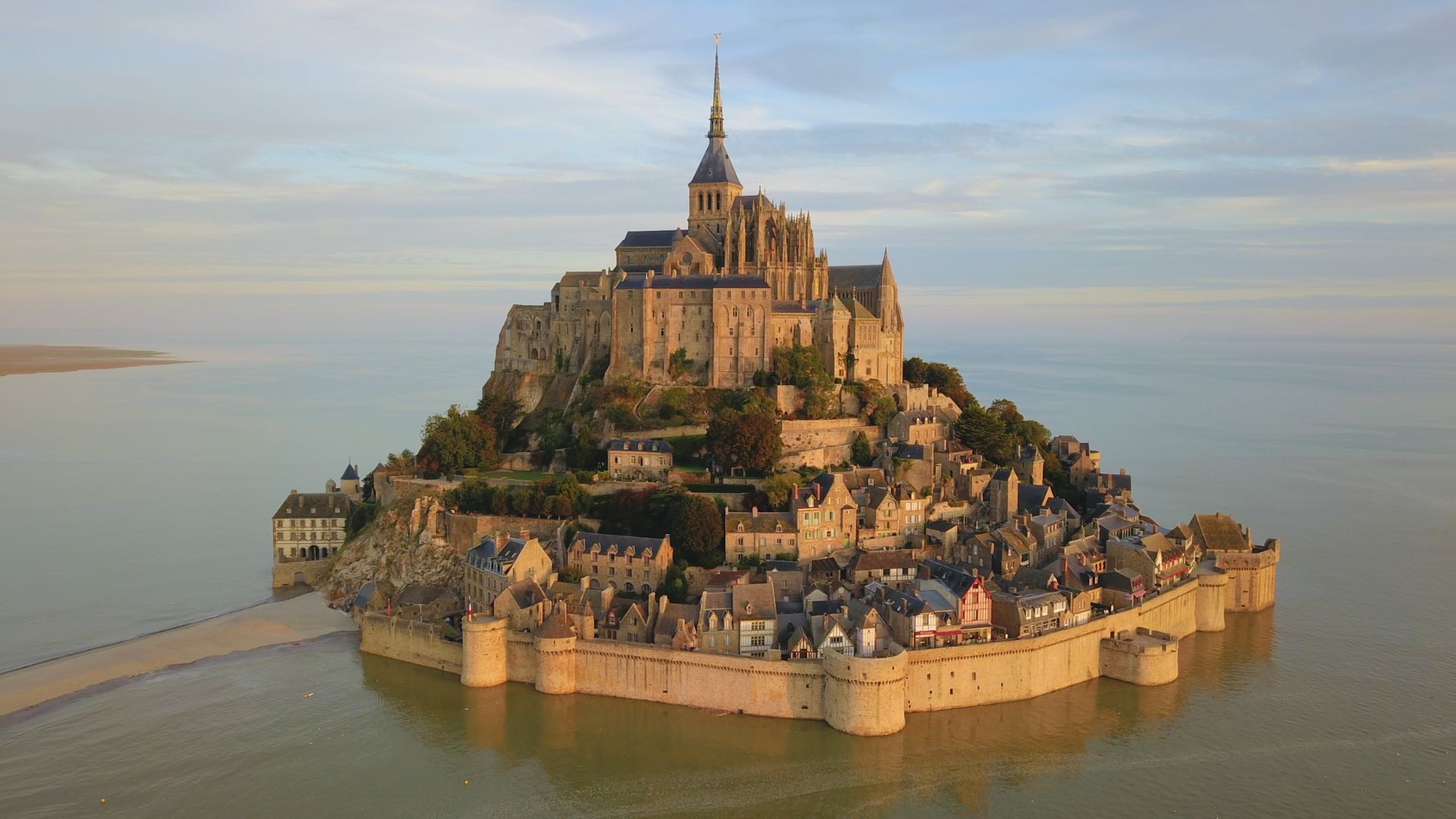 Aerial view of Mont Saint-Michel, a historic island commune in France, with a towering abbey, medieval stone structures, and serene surrounding waters.
