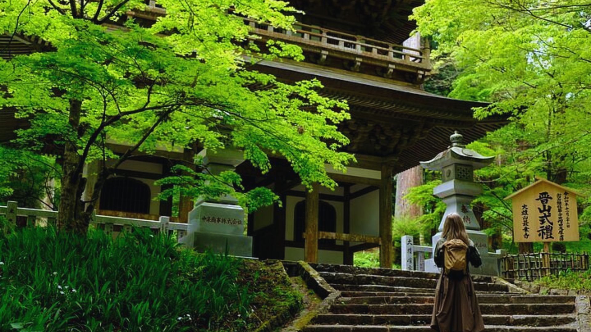 A person with a backpack stands facing a traditional Japanese temple surrounded by lush green trees. The scene is serene and inviting.