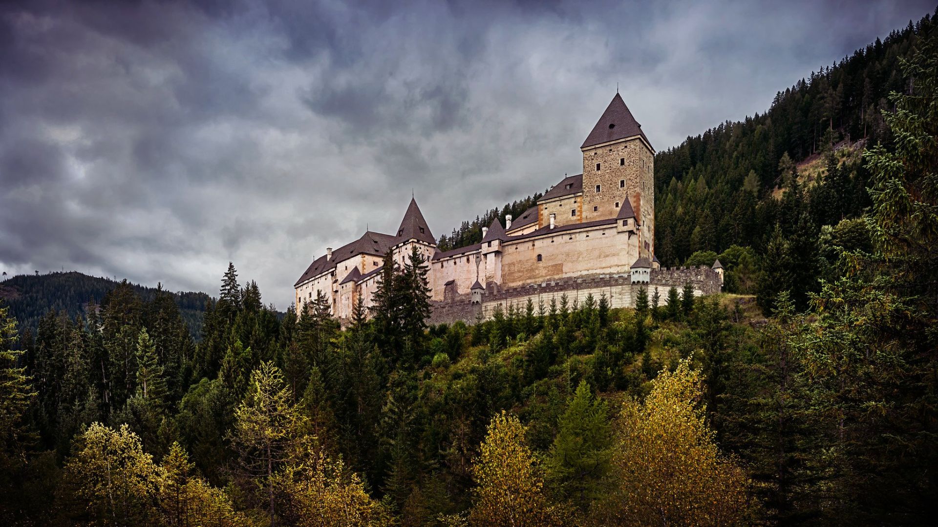 A medieval castle with tall towers sits atop a forested hill under a dramatic, cloudy sky, evoking a mysterious and historic atmosphere.