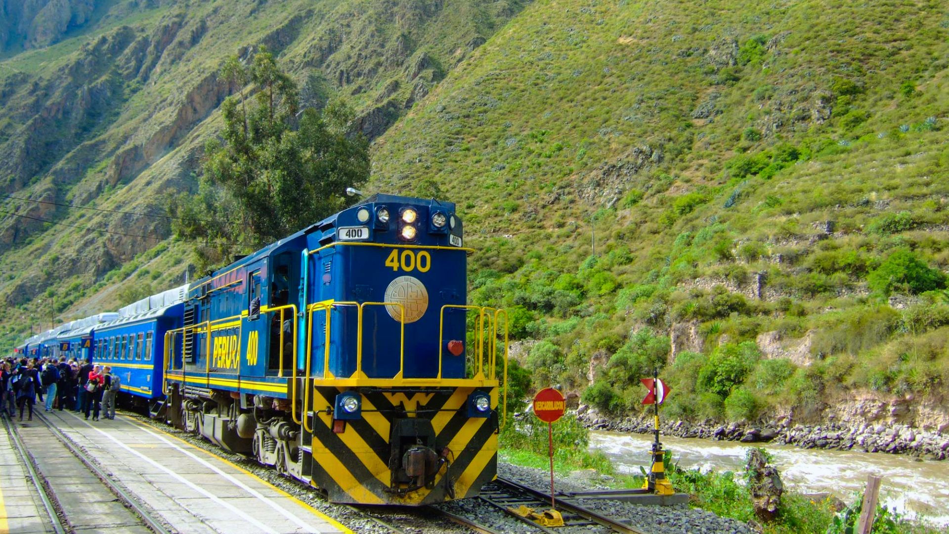 A blue and yellow train labeled "400" passes through a lush, mountainous landscape. People gather at the station, suggesting a lively, scenic journey.