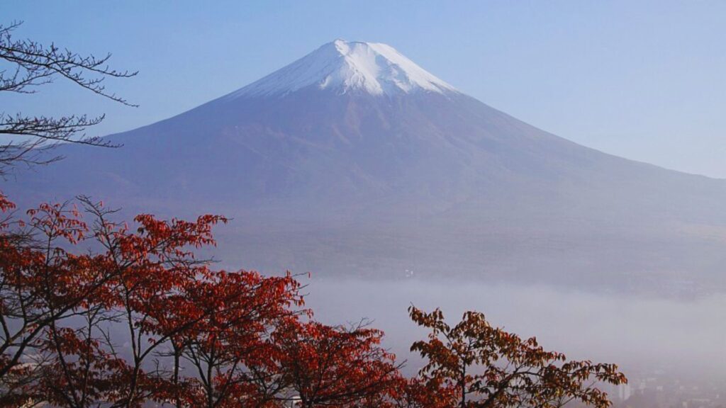 Snow-capped Mount Fuji rises majestically against a clear blue sky, framed by vibrant red and orange autumn leaves, conveying tranquility and natural beauty.