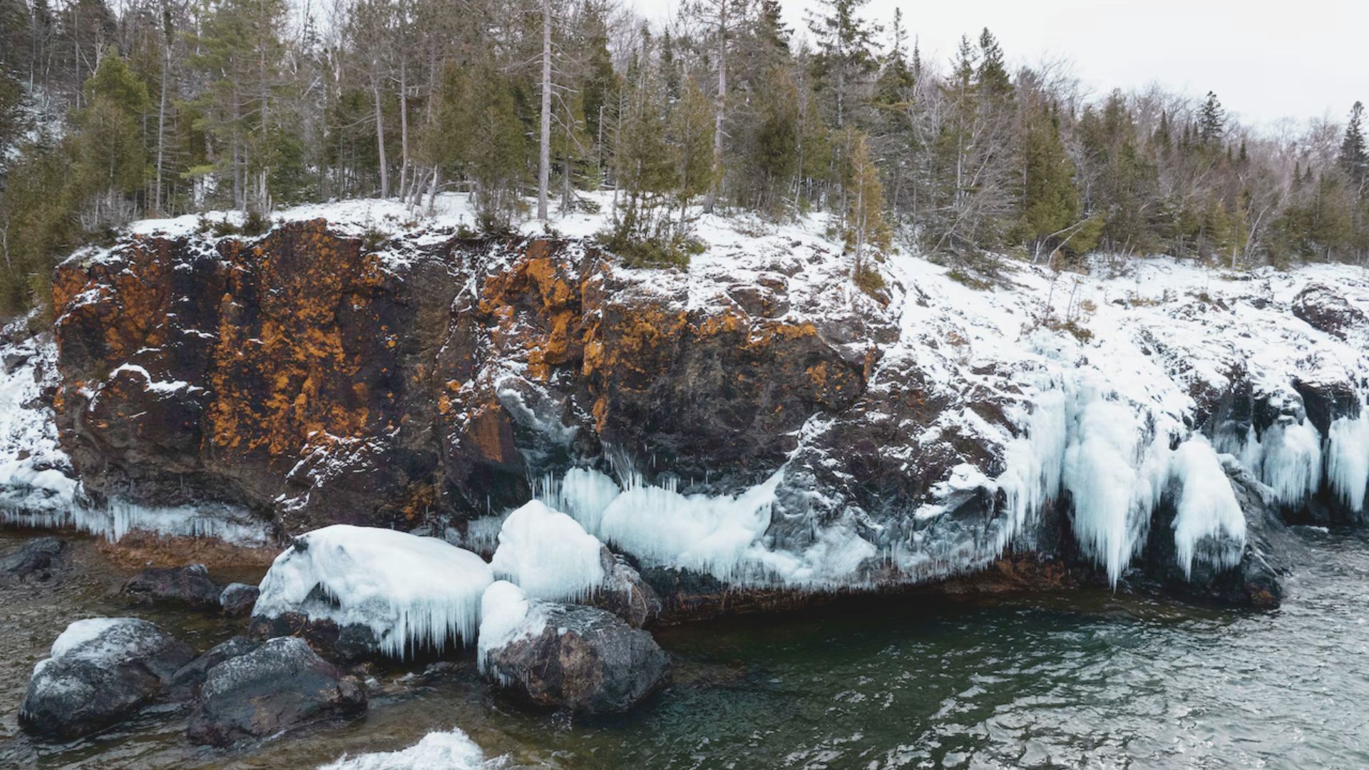 Snow-dusted rocky cliffs with lush green trees form a coastal scene. Icicles hang over the edge, meeting the clear, cold water below.