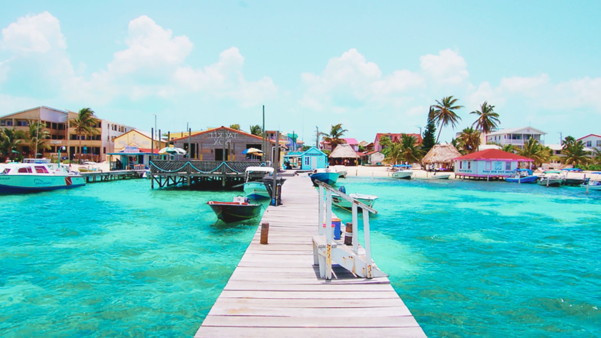 A vibrant seaside scene with turquoise waters and a wooden pier leading to a quaint, colorful village. Boats surround the pier, with palm trees in the background.