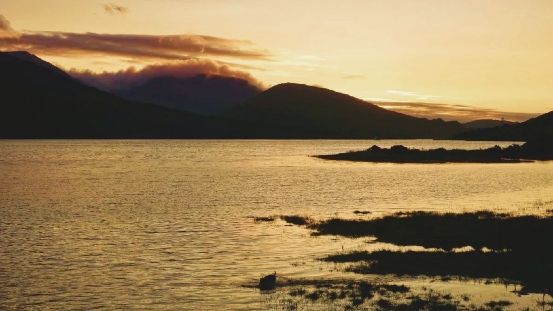 Sunset over a tranquil lake with dark silhouetted hills. Warm golden light reflects on the water, creating a serene and peaceful atmosphere.