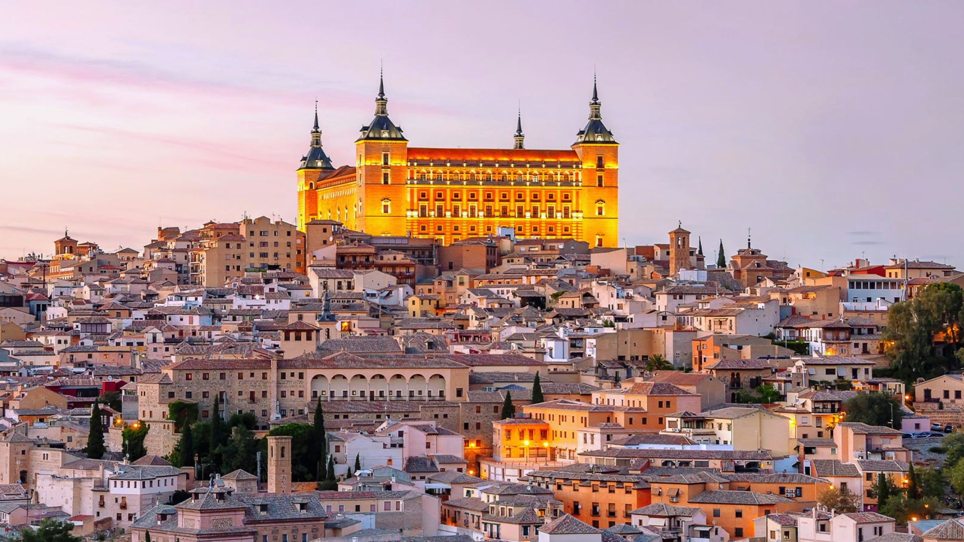 Aerial view of Toledo, Spain, at sunset. The historic Alcázar glows warmly on the hilltop, surrounded by a dense cluster of aged, tiled-roof buildings.