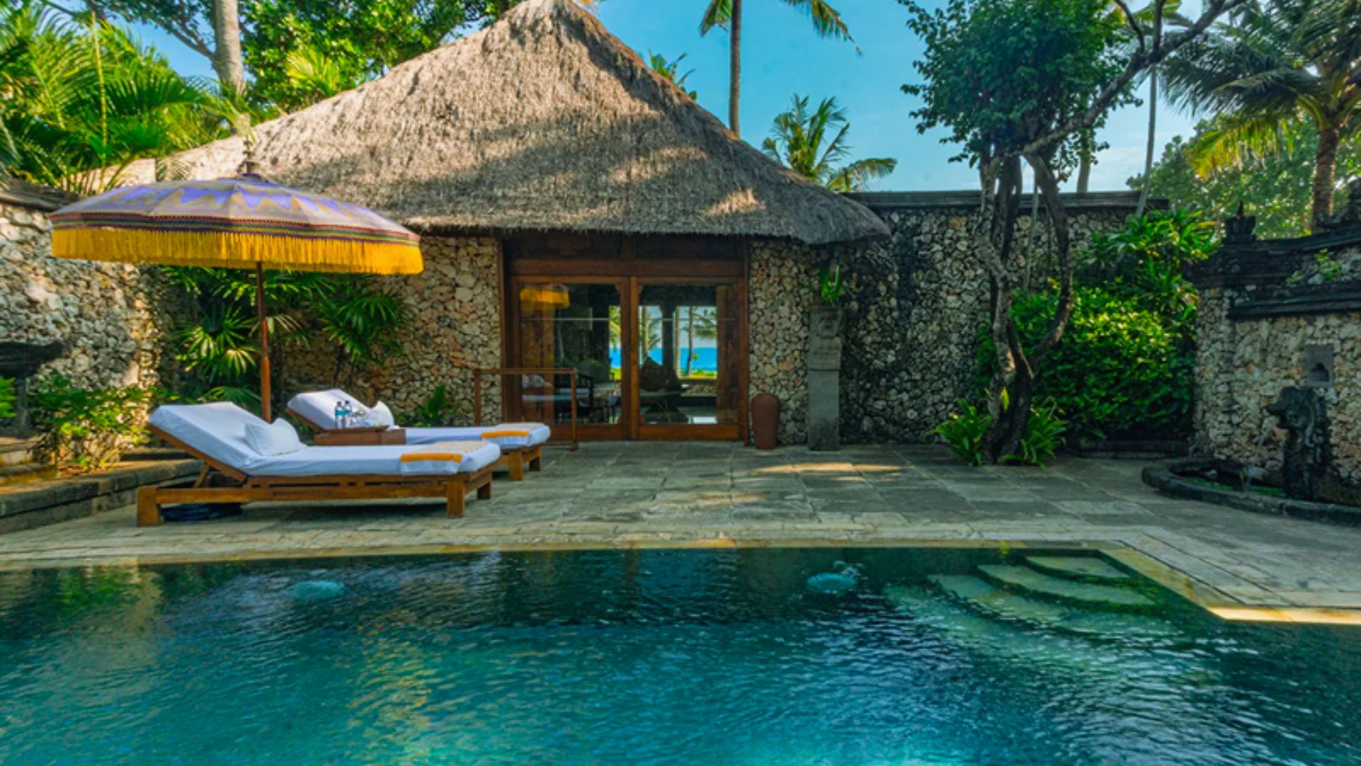 A tropical villa with a stone facade and thatched roof, surrounded by lush greenery. A pool in the foreground, with loungers and a colorful umbrella nearby.