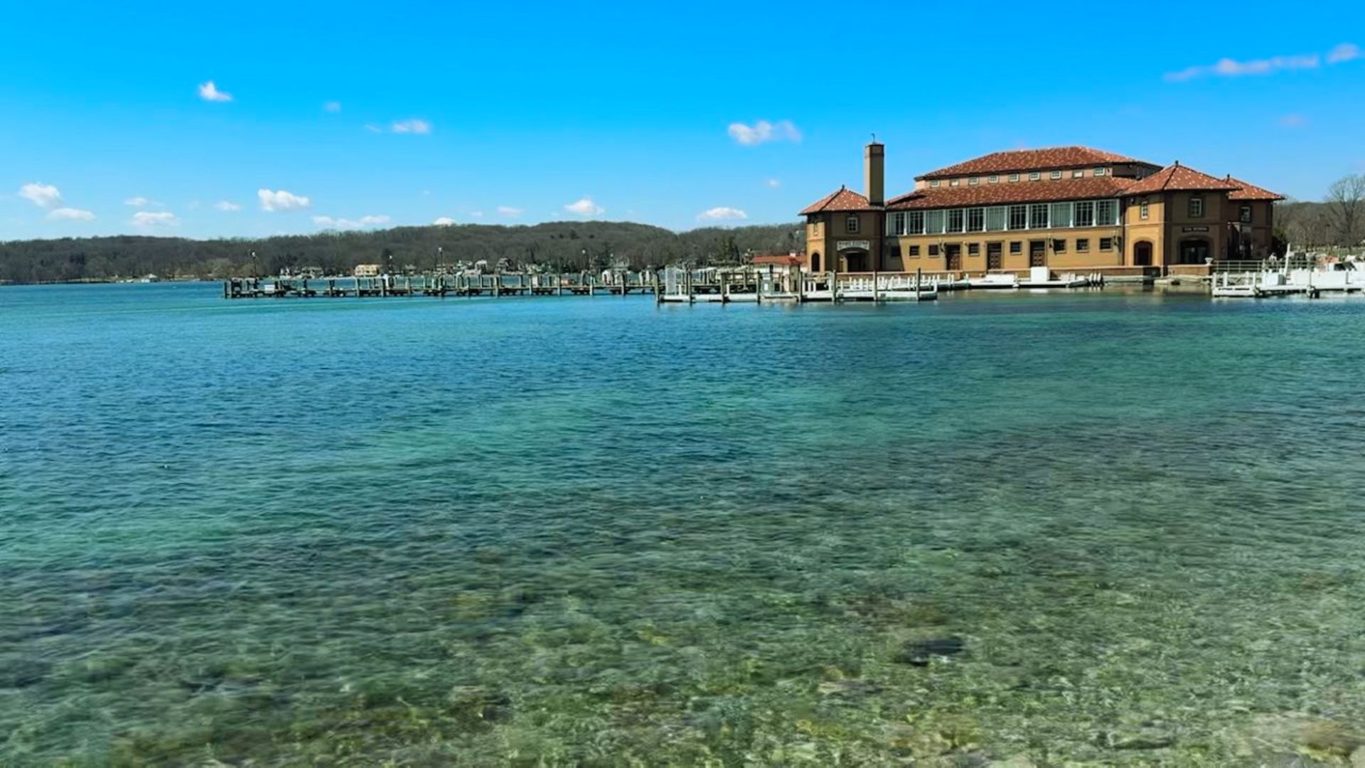 Clear blue water stretches to a building with red roofs and large windows beside a dock. The sky is bright blue with scattered clouds, creating a serene setting.