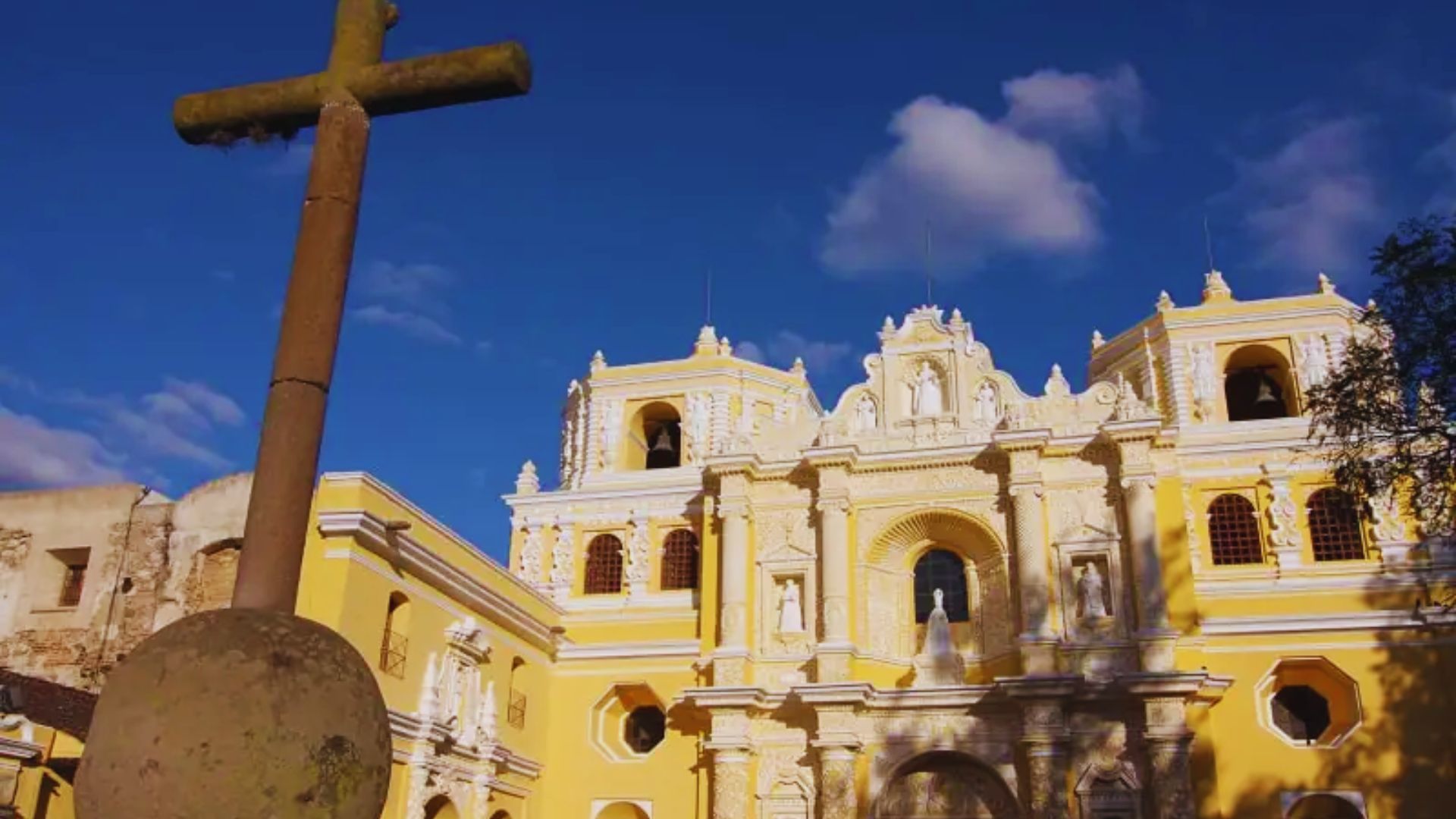 A historic yellow church with intricate white detailing is set against a blue sky. A weathered stone cross stands in the foreground, casting a serene tone.
