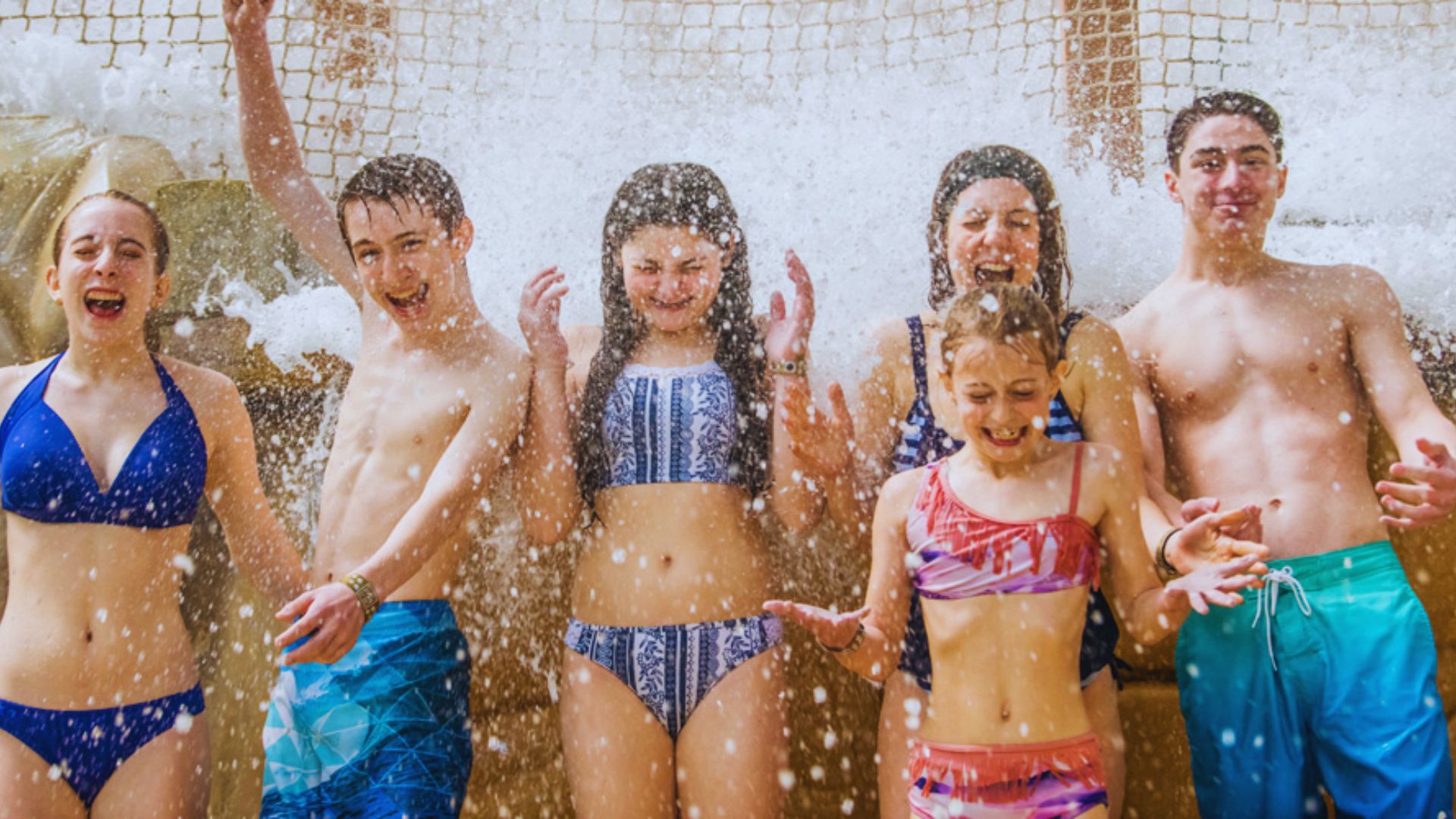 Six smiling people in swimsuits enjoy splashes of water under a waterfall at a pool. The group appears joyful and energetic against a sunny backdrop.