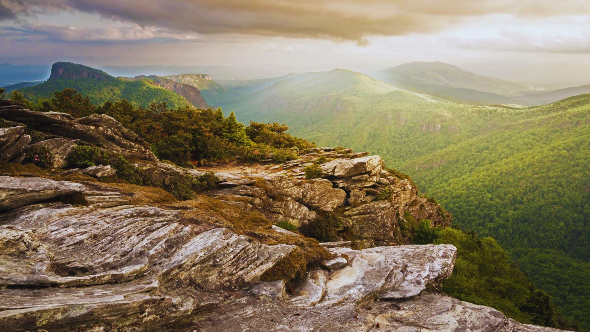 A scenic view of a rocky mountain ledge overlooking a lush, green valley. The sky is moody with clouds, and soft sunlight highlights the distant peaks.