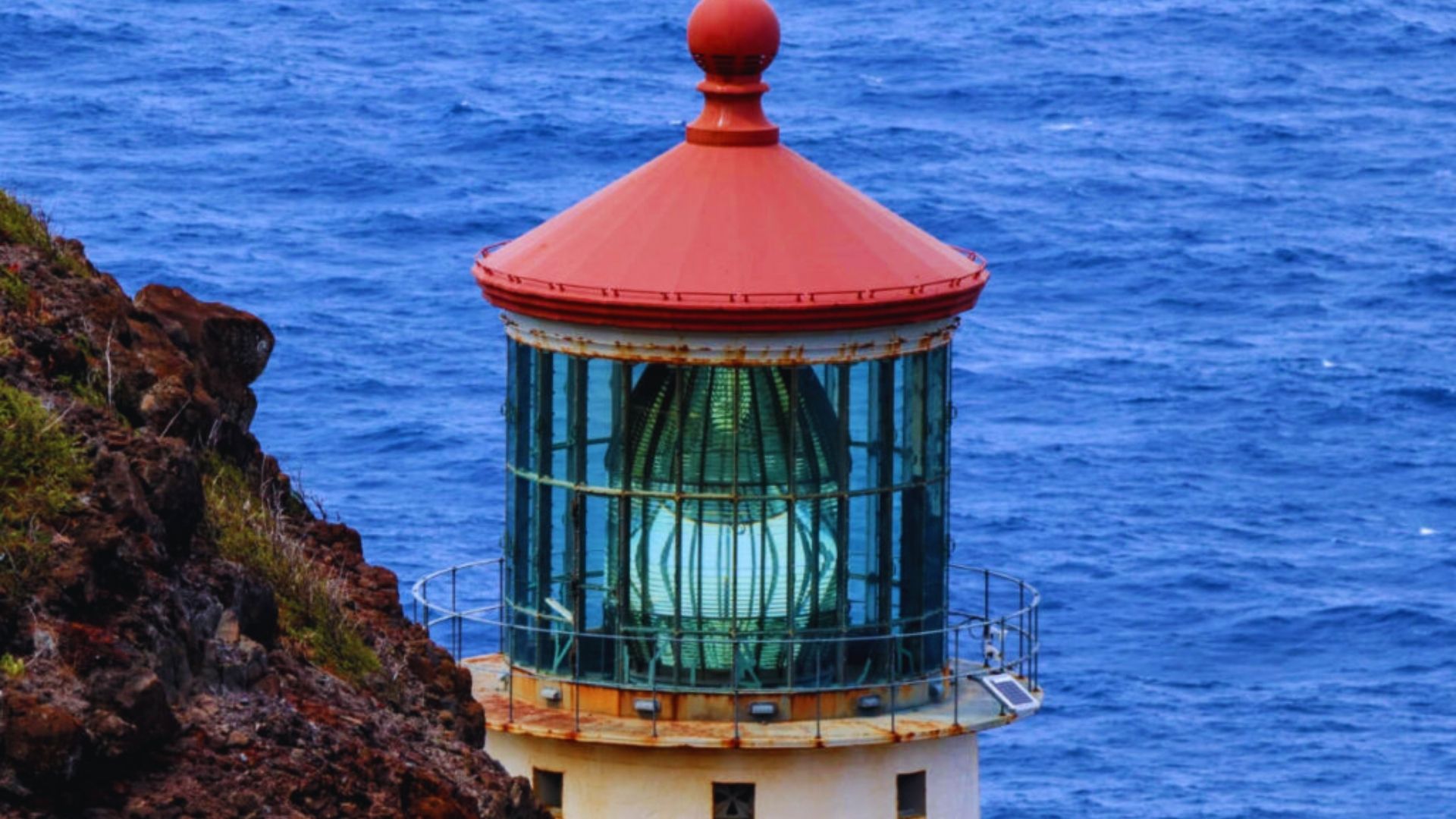 A lighthouse with a red roof stands on a rocky cliff overlooking the vast blue ocean. The glass enclosure houses a glowing light, emitting a sense of guidance.