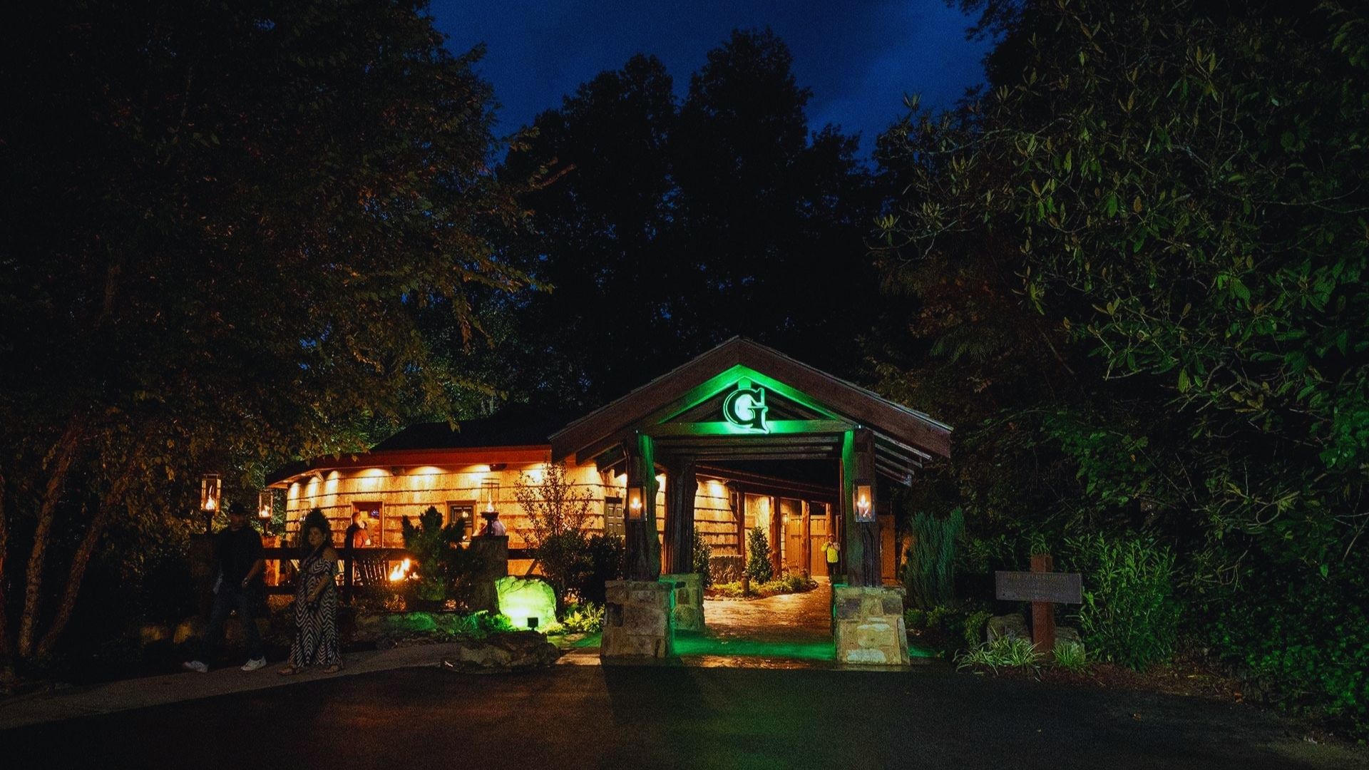 Wooden lodge warmly lit against a night sky, surrounded by trees. The entrance features a green "G" and two people stand on the path, creating a cozy atmosphere.