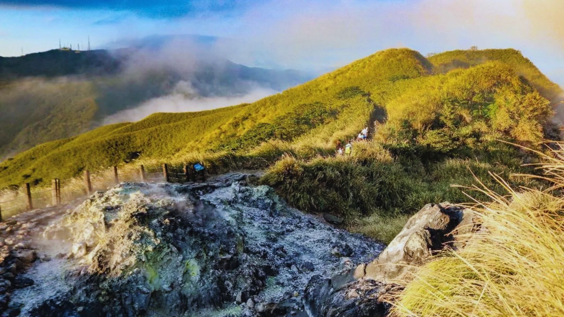 Grassy hills under a blue sky with rolling clouds. A rocky, steaming geothermal area is in the foreground. A trail with hikers winds through the landscape.