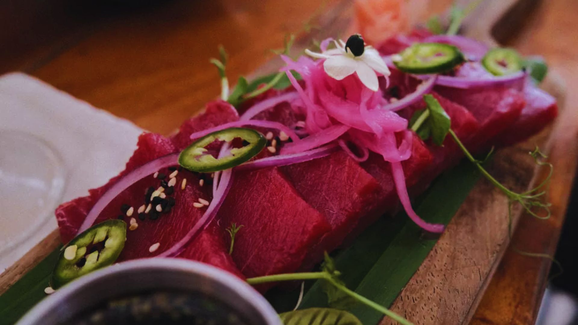 Fresh tuna sashimi with pink pickled onions, sliced jalapeños, and white flower garnish on a wooden platter, conveying elegance and freshness.