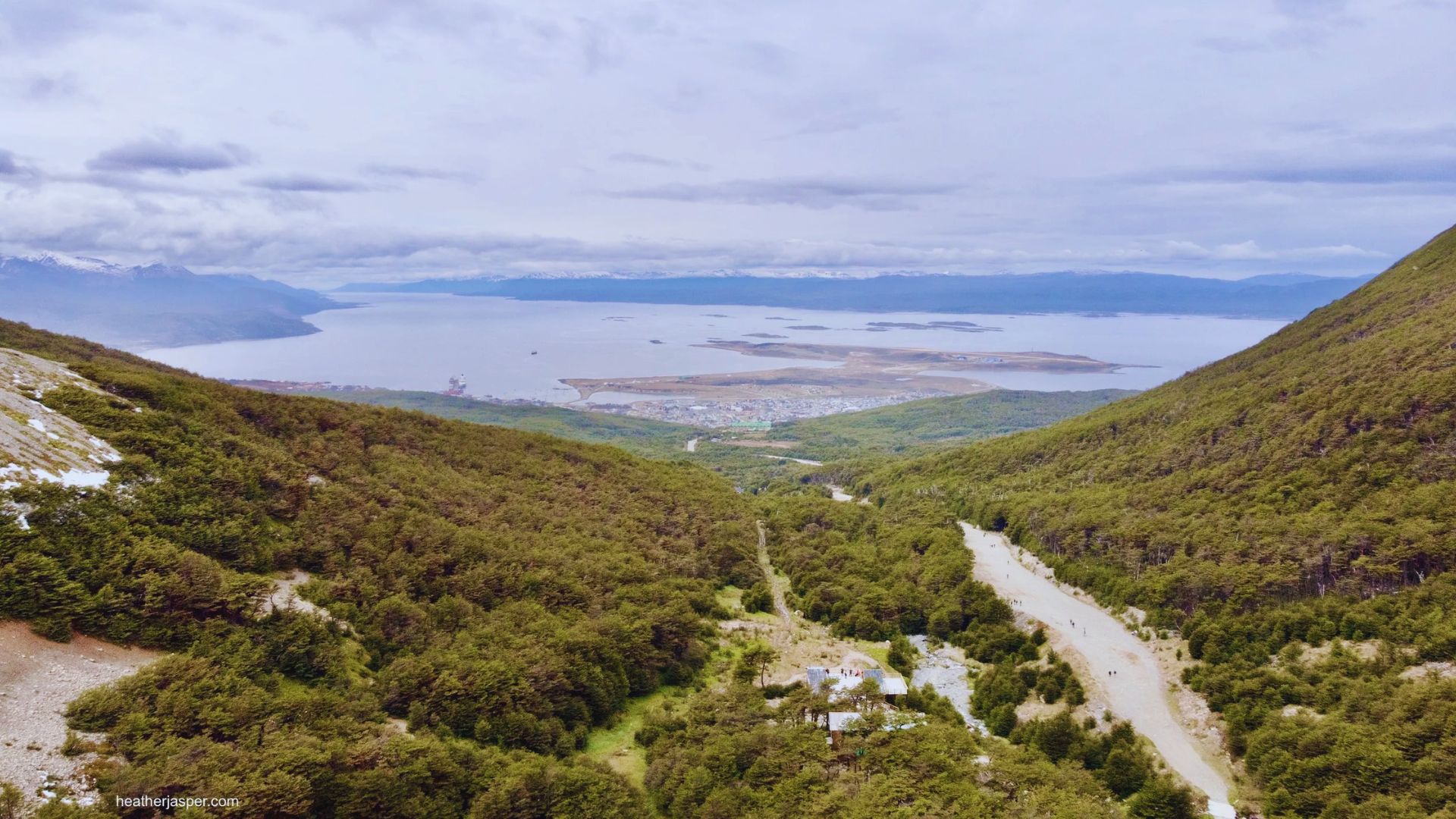 A lush green valley leads to a distant bay, with a town nestled at the water's edge. Overcast skies and rolling hills create a serene, expansive view.